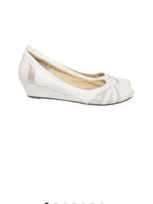Vita ballerinaskor med kilklack - Snygga vita ballerinaskor med låg kilklack och dekorativa detaljer i metallic på sidorna och framtill. Skorna har rundad tå och är tillverkade i ett slätt material som ger en stilren look. Perfekta för dig som gillar klassisk men modern stil.