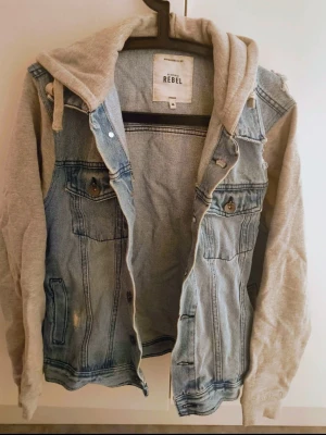 Ljusblå jeansjacka med grå hoodie - Snygg ljusblå jeansjacka från Rebel med grå sweatshirt-ärmar och huva. Jackan har slitna detaljer, knappstängning framtill och två bröstfickor. Perfekt för en avslappnad streetstil och enkel att matcha med olika outfits.