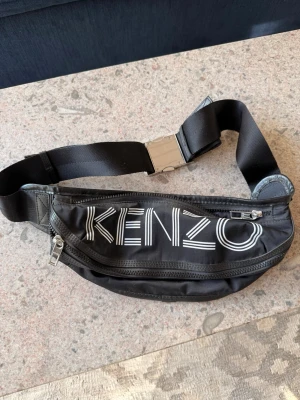 Svart magväska från Kenzo - Snygg svart magväska från Kenzo med stort vitt logotryck på framsidan. Väskan har två dragkedjefack och ett justerbart band med metallspänne. Tillverkad i slitstarkt tygmaterial, perfekt för att bära det viktigaste på ett stilrent sätt.