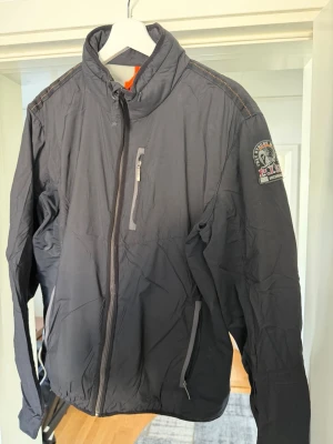 Mörkgrå vindjacka från Parajumpers - Snygg mörkgrå vindjacka från Parajumpers med dragkedja framtill, bröstficka med dragkedja och två sidofickor. Jackan har hög krage och patch med logga på ärmen. Tillverkad i lätt och slitstarkt material, perfekt för blåsiga dagar.
