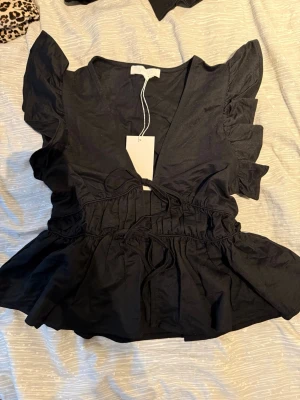 Svart volangblus från NA-KD - Supersnygg svart blus från NA-KD med volangdetaljer på ärmarna och v-ringning. Blusen har knyt i midjan och en luftig passform som ger en feminin siluett. Perfekt att styla med jeans eller kjol för en trendig look.