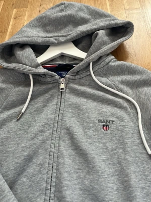 Grå zip hoodie från GANT - Snygg grå zip hoodie från GANT med broderad logga och sköld på bröstet. Hoodien har dragsko i huvan, mjukt material och ribbade muddar. Perfekt för en chill och avslappnad stil.        Nypris ca 1600kr!! MITT PRIS 650kr! (Pris går att diskutera!)