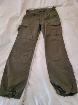 Olivgröna cargobyxor med dragsko - Säljer ett par olivgröna cargobyxor med flera stora fickor på sidorna och baktill. Byxorna har dragsko i bensluten och förstärkta knän för extra slitstyrka. Perfekta för dig som gillar en avslappnad och funktionell stil.