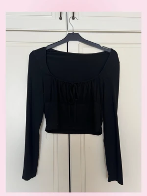 Svart croppad topp med snörning - 💗Snygg svart croppad topp med långa ärmar och en djup, rundad urringning. Toppen har en söt snörningsdetalj framtill som ger extra stilpoäng. Perfekt att matcha med höga jeans eller kjol för en trendig look. Väldigt stretchig och mjuk !💗