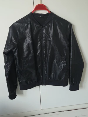 Svart glansig bomberjacka - Snygg svart bomberjacka med glansig finish och klassisk rak passform. Jackan har ribbade muddar vid ärmslut och midja samt dragkedja framtill. Perfekt för dig som vill ha en stilren och trendig look. Två öppna sido-fickor.