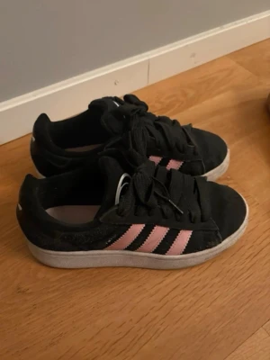Svarta Adidas sneakers med rosa ränder - Snygga svarta Adidas sneakers med klassiska tre rosa ränder på sidan och vit sula. Skorna har lågt skaft och är tillverkade i mockaliknande material med textildetaljer. Perfekta för en sportig och trendig look. Betalning genom vanlig swish! 
