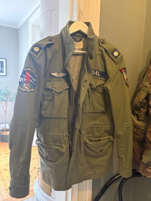 Grön field jacket från Polo Ralph Lauren - Snygg militärinspirerad grön fieldjacket från Polo Ralph Lauren med flera coola patchar och emblem på ärmar och bröst. Jackan har fyra stora fickor framtill, axelklaffar och dragkedja med knappslå. Tillverkad i slitstark bomull. Ny pris ligger den på 6000 kr. Condition 9,5/10. Storlek S men passar M bra. Vid frågor kontakta mig. 😀