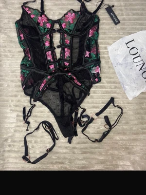 Svart spetsbody med blommor från Lounge - Säljer en svart body i mesh och spets från Lounge med broderade rosa och gröna blommor. Bodyn har justerbara axelband, dekorativa banddetaljer och medföljande strumpebandshållare. Perfekt för dig som gillar unika och snygga underkläder.