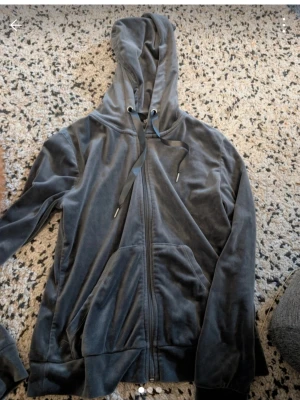 Mörkgrå velour hoodie med dragkedja stl xs - Mjuk och skön mörkgrå hoodie i velour med dragkedja framtill och stora fickor. Huva med svarta snören och ribbade muddar vid ärmslut och nederkant. Perfekt för en chill och avslappnad stil.