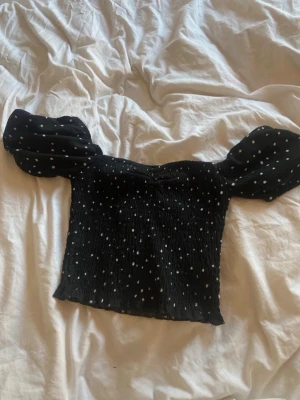 Svart prickig blus med puffärmar - Säljer en svart blus med vita prickar och stora puffärmar. Toppen har en smockad, tight passform och är tillverkad i ett lätt och stretchigt material. Perfekt för dig som gillar trendiga detaljer och en feminin look.