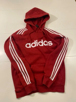 Adidas Hoodie Röd Storlek S - Röd Adidas hoodie med vit logga framtill och tre vita ränder längs ärmarna.   Storlek: S Längd: 59 cm  Armhålsbredd: 53 cm   Ställ gärna frågor! :)