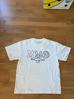 Vit Maison Margiela t-shirt - Snygg vit t-shirt från Maison Margiela med stor svart logga och texten 'MM10 Maison Margiela PARIS' på bröstet. Klassisk rund hals och rak passform. Perfekt för dig som gillar stilrena och trendiga plagg.