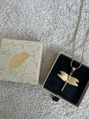 Guldfärgat ioaku halsband med trollslända - Ioaku halsband med en guldfärgad trollsländehänge. Kedjan är lite tjockare, och hänget har fina detaljerade vingar som ger ett elegant intryck. Trollsländan har tappat lite färg på baksidan men syns inte. 