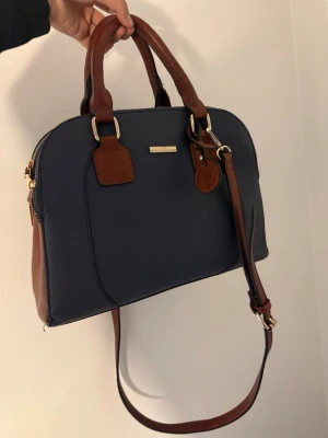 Mörkblå handväska med bruna detaljer - Snygg mörkblå handväska med bruna handtag och axelrem i läder. Väskan har guldfärgade metalldetaljer och en klassisk, rundad form. Perfekt för dig som gillar stilrena accessoarer med lyxig känsla.