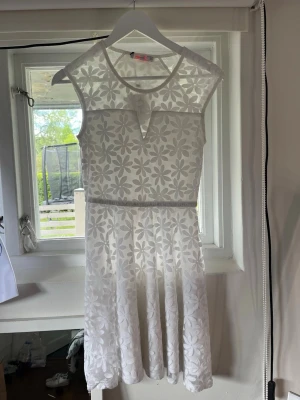 Vit blommig studentklänning med spets - Söt vit kortklänning med blommigt spetsmönster och transparenta partier upptill. Klänningen har ärmlös design, markerad midja och lätt utställd kjol. Perfekt för sommarens alla tillfällen.