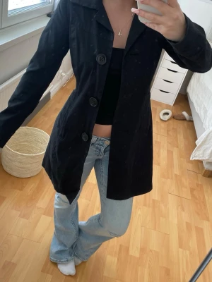 Svart klassisk rock med stora knappar - Snygg svart rock med tre stora knappar framtill och klassisk krage. Rak modell som går ner över höfterna, perfekt att slänga över en outfit för en stilren look. 