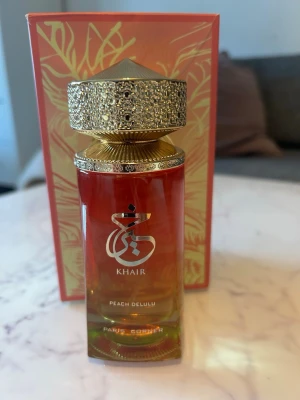 Khair Peach Delulu parfym - Khair Peach Delulu från Paris Corner är en parfym i en lyxig glasflaska med guldigt, detaljerat lock. Flaskan har en gradient i orange och gul med elegant logga framtill. Doften är inspirerad av persika och levereras i en snygg förpackning med guldmönster.