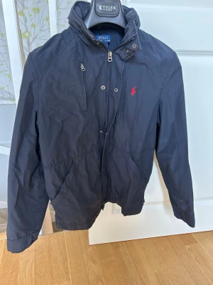 Mörk blå vindjacka från Polo Ralph Lauren - Snygg blå vindjacka från Polo Ralph Lauren med dragkedja, och knappar med röd logga på bröstet. Jackan har praktiska fickor och är tillverkad i ett lätt och slitstarkt material. Luva som är möjlig att hålla infälld. Är använd men i gott till mkt gott skick. Strl L (14-16)