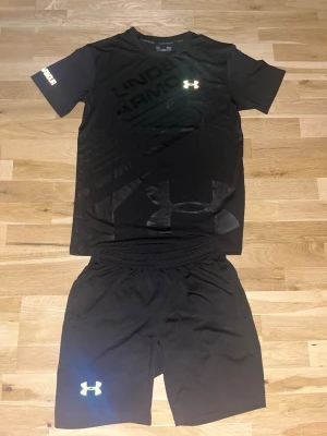 Svart träningsset från Under Armour - Storleken är mellan S-M.    Jag Säljer ett svart träningsset från Under Armour med t-shirt och shorts. Setet har reflekterande loggor på både tröjan och shortsen samt ett coolt holografiskt tryck på shortsen. Perfekt för träning och sportaktiviteter eller bara ha på Sommaren!