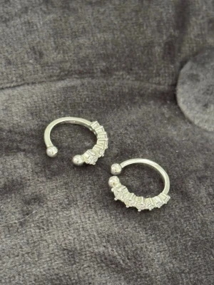 Silvriga örhängen med pärldetaljer - Silvriga örhängen i halvöppen ringdesign med små pärlliknande detaljer längs ena sidan. De är till för att sätta fast på öronen där man inte har egna hål. 