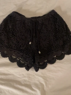 Svarta spetsiga shorts med volang - Snygga svarta shorts från am spets och volangkant. De har ett detaljerat blommigt mönster i spetsen och små dekorativa pärlor framtill. Perfekta för dig som vill ha en feminin och trendig look.