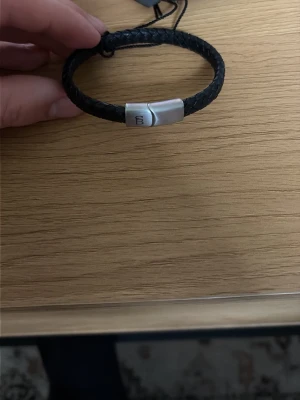 Svart flätat armband Steel & Barnett - Snyggt svart flätat armband från Steel & Barnett med ett stilrent magnetlås i silverfärgad metall. Armbandet är gjort av läder och har en modern, minimalistisk design som passar perfekt till din vardagsstil.