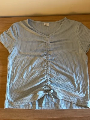 Ljusblå ribbad topp med knyt - Snygg ljusblå ribbad topp från GINA TRICOT med kort ärm, v-ringning och rynkdetalj framtill som avslutas med en knytning. Perfekt för en trendig och avslappnad look.