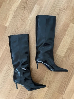 Svarta knähöga stövlar - Säljer ett par svarta knähöga stövlar från H&M i glansigt lackmaterial med spetsig tå och smal klack. Snygg och stilren design som ger en edgy vibe till outfiten. Perfekta för dig som vill sticka ut och addera lite attitude till din look🖤