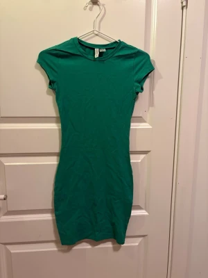 Grön kortärmad klänning H&M Divided - En enkel och snygg grön klänning från H&M Divided. Modellen är kortärmad och har en kroppsnära passform med rund hals. Perfekt till sommaren och lätt att styla med accessoarer. Tillverkad i mjuk bomull för skön känsla. Jag har tidigare haft katter så katthår kan förekomma men kläderna är alltid tvättade innan de skickas. 