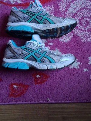 Asics vita och turkosa sneakers - Säljer ett par snygga Asics sneakers i vitt, grått och turkost med svarta detaljer. Skorna har meshpaneler för bra ventilation och en stabil sula med GEL-dämpning. Perfekta för dig som gillar sportig stil och vill ha bekväma sneakers med cool design i storlek 39
