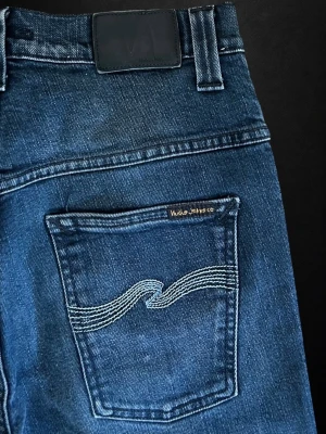 Nudie jeans - Snygga blå jeans från Nudie Jeans Co med klassisk femficksdesign och ikonisk vågstickning på bakfickorna. Jeansen har rak passform och är tillverkade i slitstark denim med en cool tvättad look. Perfekta för en avslappnad och trendig stil.