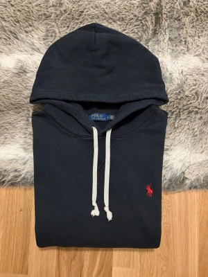 Svart Polo Ralph Lauren Hoodie - Storlek L - Hej! 👋 Säljer en riktigt snygg svart Polo Ralph Lauren hoodie i storlek L med röd pony-logga. 🐎 Tröjan är i mycket fint skick, helt utan hål, fläckar eller andra defekter. Välvårdad och fräsch! 100% äkta med originaltvättlappar kvar (ingen QR-kod på just denna årsmodell). Ett klockrent premium-basplagg som passar till allt och håller stilen. Fraktar snabbt! 💬 🚛📦💯 