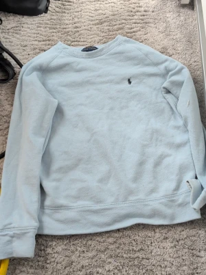 Ljusblå sweatshirt från Polo Ralph Lauren - Ljusblå sweatshirt från Polo Ralph Lauren med klassisk rund hals och diskret broderad logga på bröstet. Tröjan har långa ärmar och är tillverkad i mjukt bomullsmaterial som känns skönt mot huden. Perfekt för en avslappnad och stilren look.