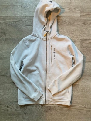Vit hoodie från Sail Racing - Vit hoodie från Sail Racing med dragkedja framtill och en bröstficka med logga. Klassisk modell med huva och långa ärmar. Perfekt för en avslappnad och sportig stil. Mjukt material som känns skönt mot huden.