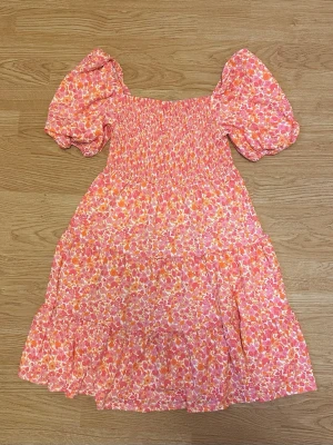 Blommig rosa orange klänning med puffärm midsommar sommar - Klänning med blommigt mönster i rosa, orange och vitt. Modellen har smockad överdel och puffiga korta ärmar