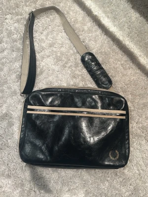 Svart axelväska från Fred Perry - Snygg svart axelväska från Fred Perry i glansigt konstläder med två beige ränder på framsidan och klassisk logga i hörnet. Väskan har justerbar axelrem i beige och svart med vadderad axelplatta. Perfekt för att bära dina grejer med stil.