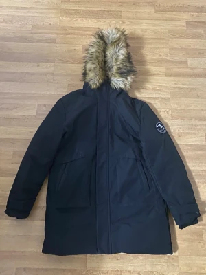 Svart parkas med fuskpäls från Everest - Svart parkas med huva och avtagbar fuskpäls. Jackan har två stora fickor framtill, dragkedja och tryckknappar. Fodrad insida för extra värme och patch med logga på ärmen. Perfekt för kalla dagar.