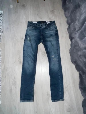 Blå slim fit jeans från Jack Jones  - Säljer ett par blå slim fit jeans från Jack & Jones med snygga slitningar och tvättade detaljer. Jeansen har klassisk femficksmodell, normal midja och dragkedjegylf. Perfekta för en avslappnad och trendig look. 