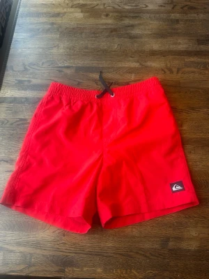 Röda badbyxor från Quiksilver juniormodell  - Snygga röda badbyxor från Quiksilver med svart dragsko i midjan och klassisk logga på ena benet. Tillverkade i snabbtorkande material och har innershorts i mesh för extra komfort. Perfekta för stranden eller poolen.
