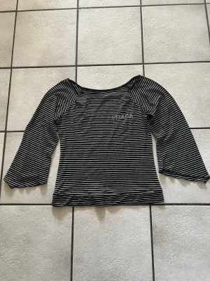 Randig svartvit topp storlek M - Snygg långärmad topp med svartvitt randigt mönster och broderad logga på bröstet. Toppen har en något vid halsringning och är tillverkad i mjukt material som känns skönt mot huden. Perfekt för dig som gillar en enkel men trendig stil.