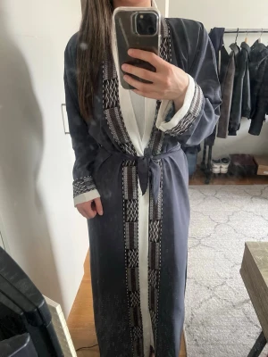 Svart lång koftan (abaya) med mönster - Köpt Utomlands. Fin detaljerad koftan klänning (abaya) i storlek xxl som passar mig (har vanligtvis s). Man kan knyta eller ha den öppen.