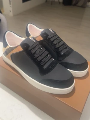 Burberry svarta sneakers med rutigt mönster - Snygga svarta sneakers från Burberry med klassiskt beige, svart och vitt rutmönster på hälen. Skorna har svarta snören, vit sula och detaljer i mocka och textil. Perfekta för dig som vill ha en stilren och lyxig look med en touch av ikoniskt mönster.