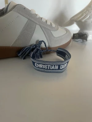 Blått Christian Dior tygarmband - Snyggt blått tygarmband från Christian Dior med vit brodyr och texten 'CHRISTIAN DIOR J'ADIOR'. Armbandet har justerbar knytning med tofsar i ändarna och en cool, avslappnad stil. Perfekt accessoar för att lyfta din outfit.