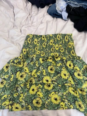 Blommig gul kjol Holly & Whyte XS - Supersöt kort kjol som också kan användas som en topp från Holly & Whyte by Lindex i storlek XS. Kjolen har ett gult blommönster med gröna blad och smock i midjan för en bekväm passform. Perfekt för soliga dagar och ger en fräsch sommarkänsla.