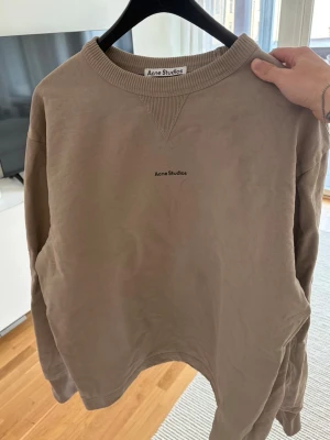 Beige sweatshirt från Acne Studios - Säljer en stilren beige sweatshirt från Acne Studios med diskret logga på bröstet. Tröjan har rund halsringning, långa ärmar och är gjord i mjukt material. Perfekt för en avslappnad och trendig look.
