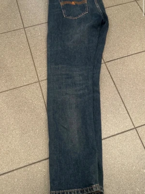 Blå raka jeans från Nudie Jeans - Säljer ett par klassiska blå jeans från Nudie Jeans med slim ben och femficksmodell. Jeansen har snygga slitningar och kontrastsömmar, samt en bakficka med Nudie-logga. Perfekta för en avslappnad och stilren look.