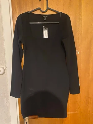 Svart långärmad klänning - En stilren svart klänning från Bik Bok med lång ärm och fyrkantig ringning. Klänningen är kort och har en tight passform, perfekt för dig som gillar en enkel men trendig look. Tillverkad i mjukt material som sitter skönt på kroppen.