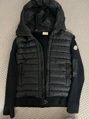 Moncler cardigan jacka  - Snygg svart pufferjacka från Moncler med vadderad front och huva. Ärmarna är ribbstickade och jackan har dragkedja framtill samt Moncler-logga på ena ärmen. Perfekt för kalla dagar och riktigt trendig stil.