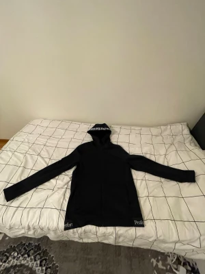 Peak Performance kofta - Svart hoodie från Peak Performance med logga på huvan och nedtill. Klassisk passform med långa ärmar och huva. Perfekt för en avslappnad och sportig stil.