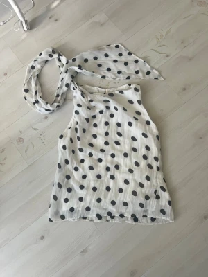 Vit prickig linnetopp från MNG - Snygg vit ärmlös linnetopp från MNG med svarta prickar och en knytbar scarf-detalj vid halsen. Perfekt för dig som gillar trendiga och lekfulla plagg. Luftigt material som passar till många olika stilar.